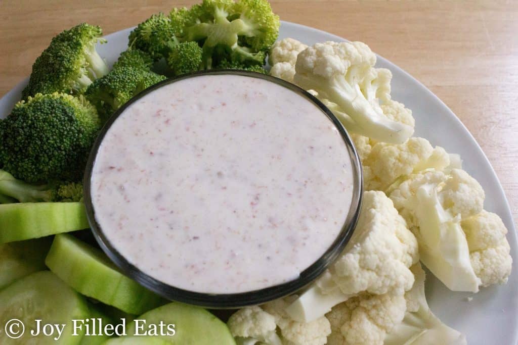 close up overhead view of a bowl filled with creamy bacon dip surrounded by a platter of vegetables including broccoli florets, cauliflower florets and cucumber slices