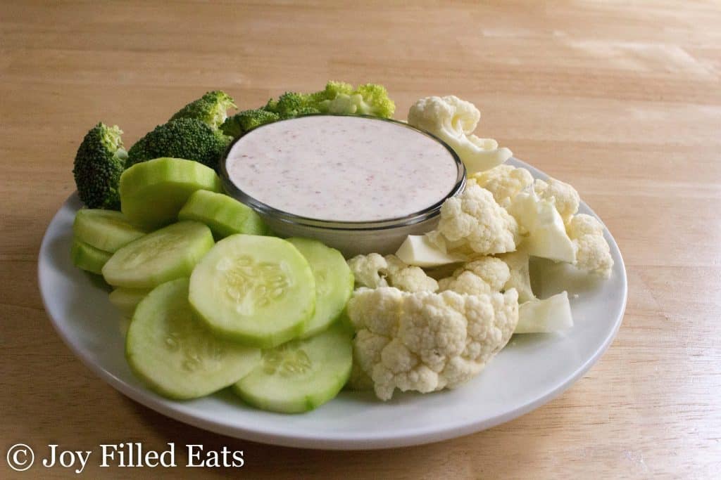 a bowl filled with creamy bacon dip surrounded by a platter of vegetables including broccoli florets, cauliflower florets and cucumber slices