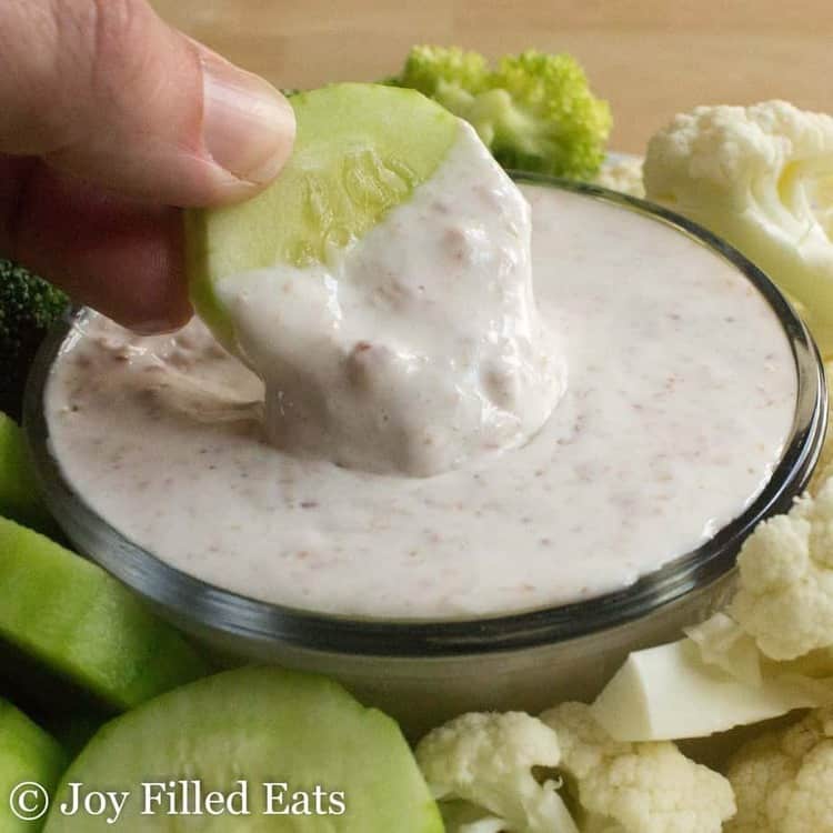 hand holding a slice of cucumber being dipped into a small bowl of creamy bacon dip