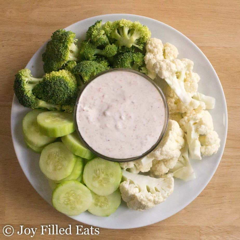 overhead view of a bowl filled with creamy bacon dip surrounded by a platter of vegetables including broccoli florets, cauliflower florets and cucumber slices