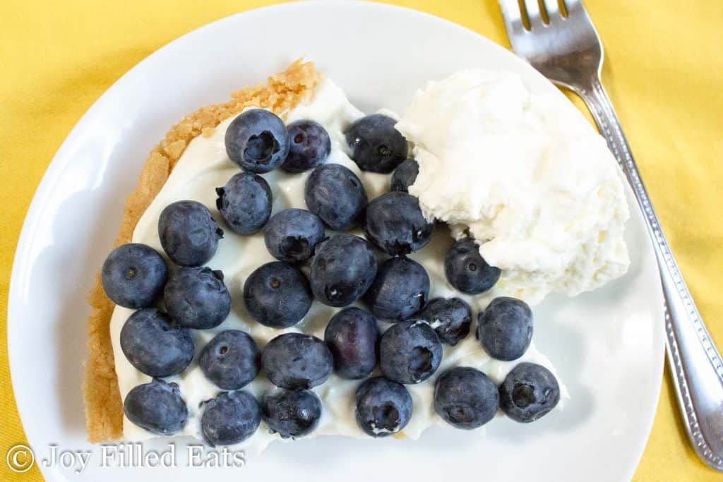 overhead view of a slice of lemon ricotta pie topped with fresh blueberries and a dollop of whipped cream on a white plate with a fork