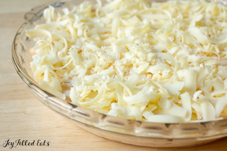 pie plate of hot cheddar onion dip ingredients topped with shredded cheese before baking