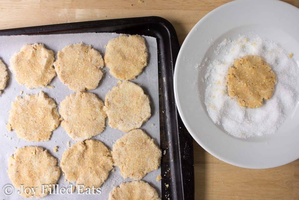 cookie dough discs lined on a sheet pan next to a bowl of sweetener with one disc placed in the sweetener