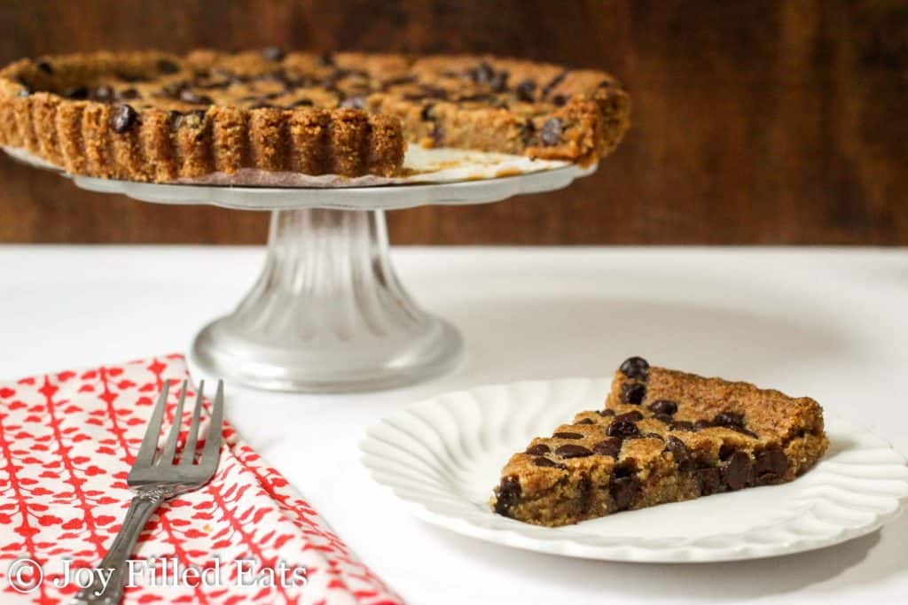 slice of chocolate chip cookie tart on a white plate next to cake tray with cookie tart on top