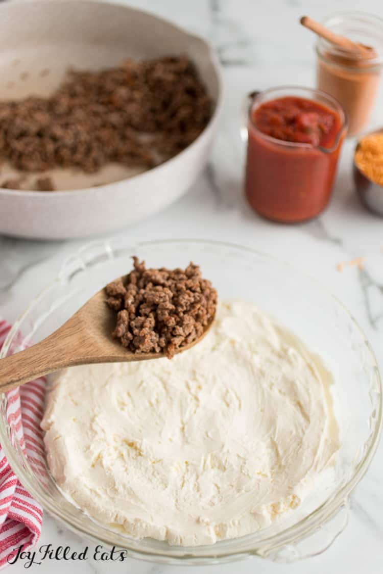 wooden spoon holding ground meat above layer of cream cheese in pie plate for taco dip