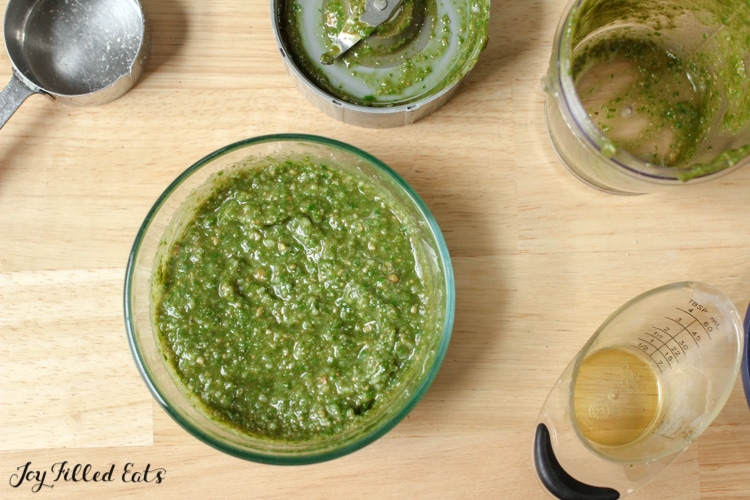 Bowl of nut free pesto surrounded by used cooking utensils including metal measuring cup, small food processor with lid and small liquid measuring cup