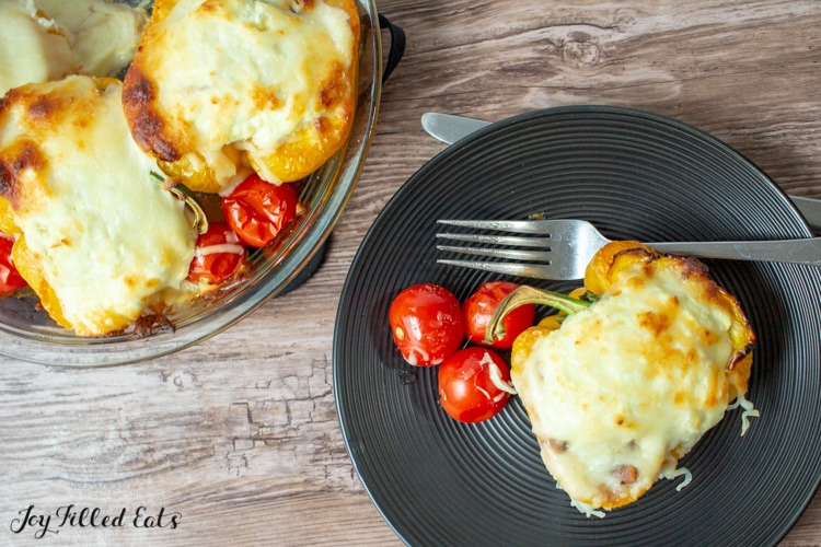 Black plate and fork with a white lasagna stuffed pepper and roasted cherry tomatoes. Next to glass pie plate with three white lasagna stuffed peppers and roasted tomatoes