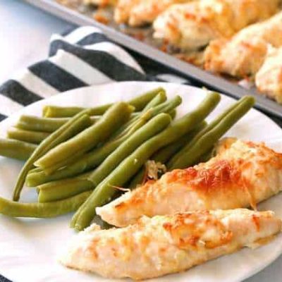plate with garlic parmesan chicken tenders and green beans set next to a sheet pan lined with garlic Parmesan chicken tenders
