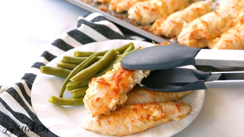 tongs placing a garlic Parmesan chicken tender onto a plate of more chicken tenders and a side of green beans