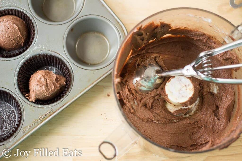 dark chocolate cupcake batter scooped into cupcake wrapper lined muffin tin next to bowl and ice cream scoop of more batter