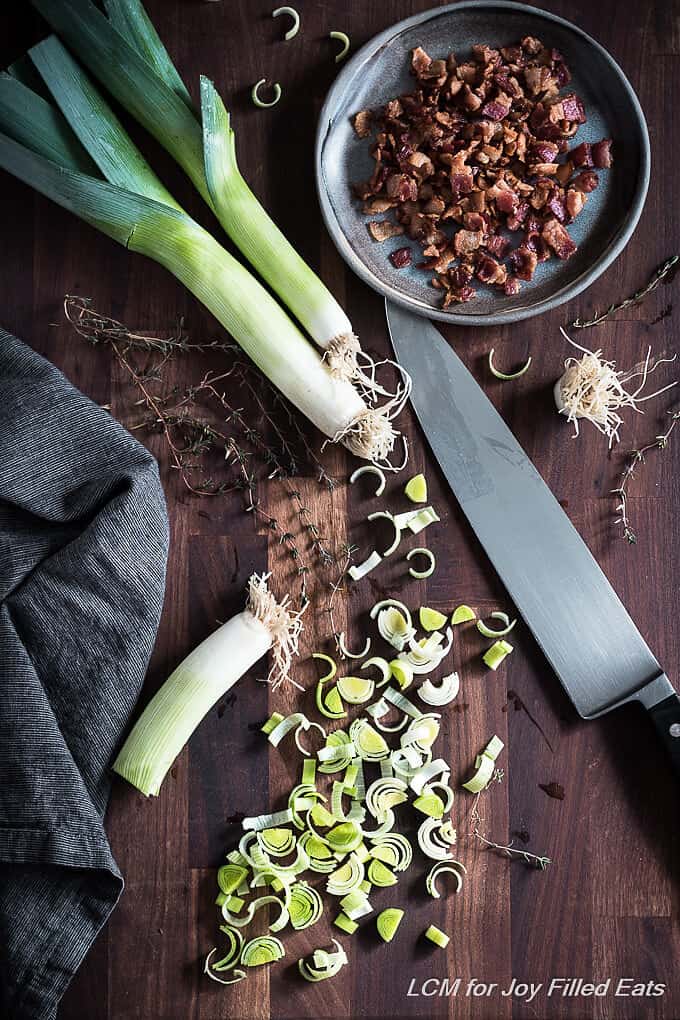 sliced leeks on a cutting board with knife and plate of crumbled bacon