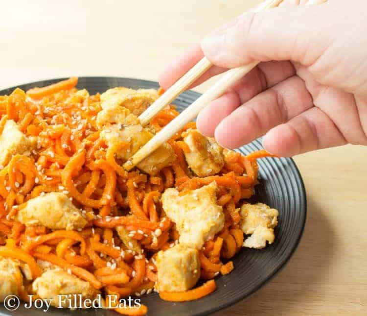 hand holding chopsticks reaching in for a bite of Sweet Ginger Chicken with Carrot Noodles on a black plate