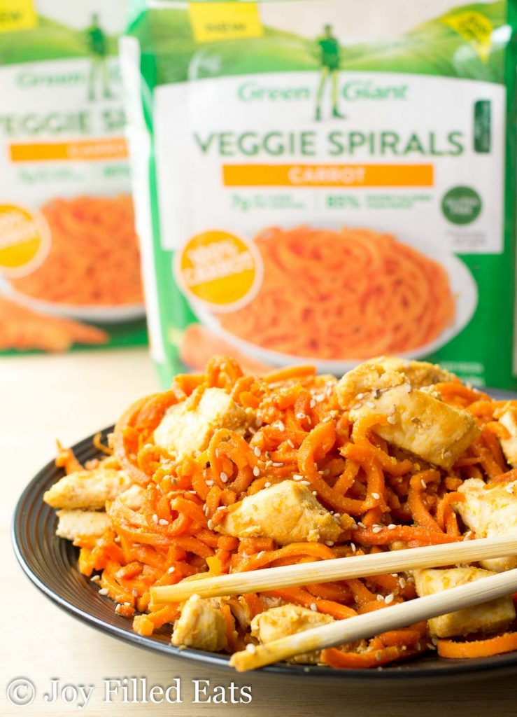 black plate of Sweet Ginger Chicken with Carrot Noodles with chopsticks resting on rim set in front of two packages of Green Giant Carrot Veggie Spirals