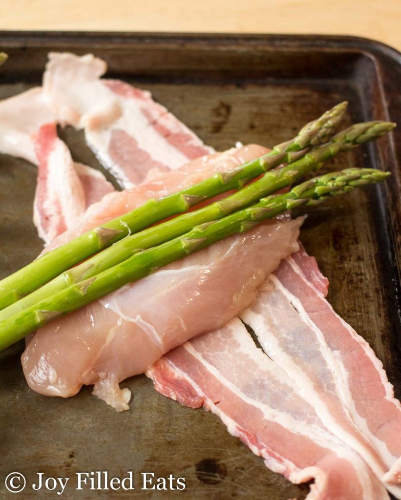 asparagus placed on top of chicken pieces laying on two strips of bacon on a sheet pan