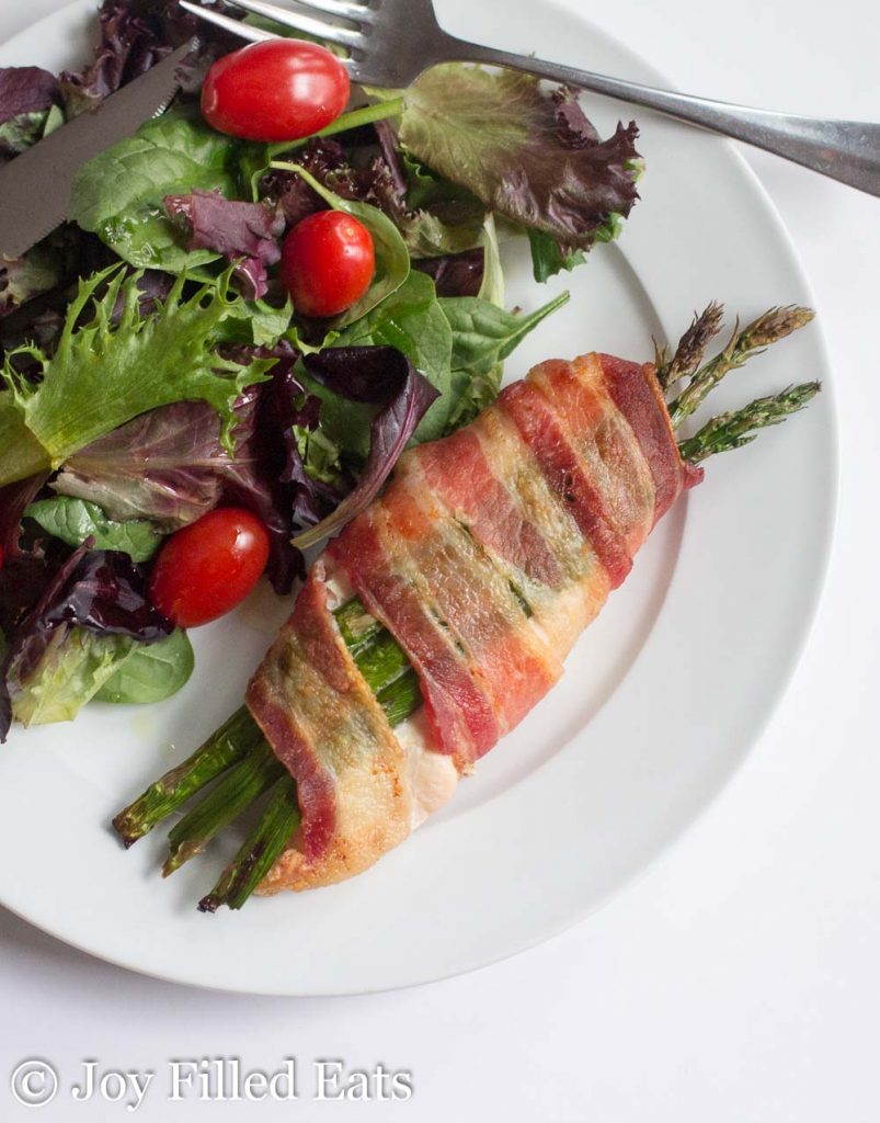 asparagus stuffed chicken wrapped in bacon on a white with a side salad and fork from above