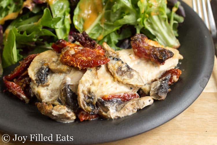 baked chicken breast with mushrooms and sun dried tomatoes on a black plate with a mixed green salad