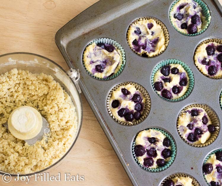 muffin tin filled with blueberry muffin batter next to a food processor with crumb topping