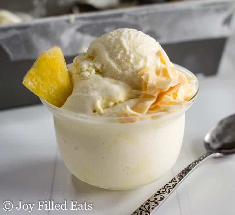 scoop of pina colada ice cream topped with coconut flakes and pineapple in a small glass bowl next to a spoon