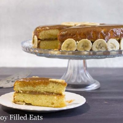 slice of banana foster cake on a white plate placed next to a cake plate with remaining banana cake