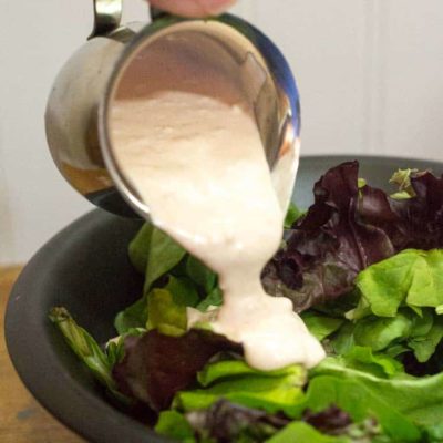 hand holding small pitcher pouring creamy garlic salad dressing onto mixed greens