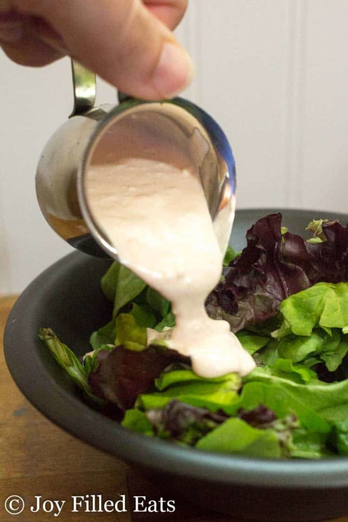 hand holding small pitcher pouring creamy garlic salad dressing onto mixed greens