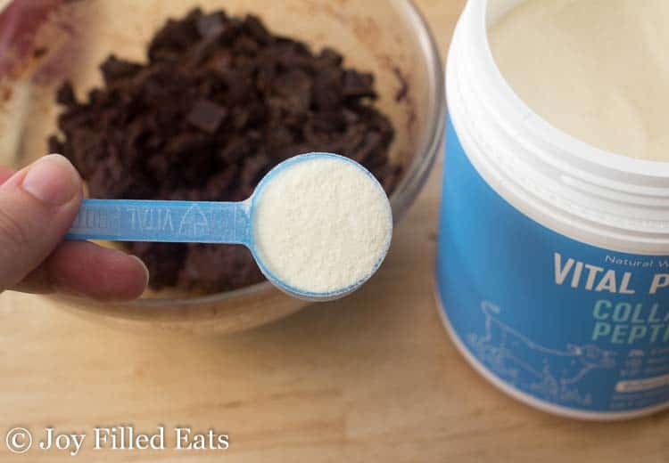 hand holding tablespoon of collagen peptide powder above a bowl of chocolate pieces