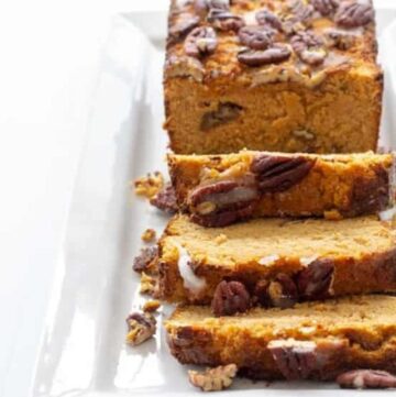 pumpkin bread loaf topped with pecans on a white platter with three slices cut from loaf