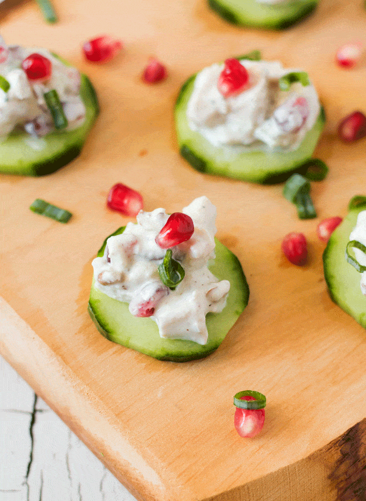 close up of pomegranate chicken salad cucumber bites on a piece of wood topped with pomegranate seeds