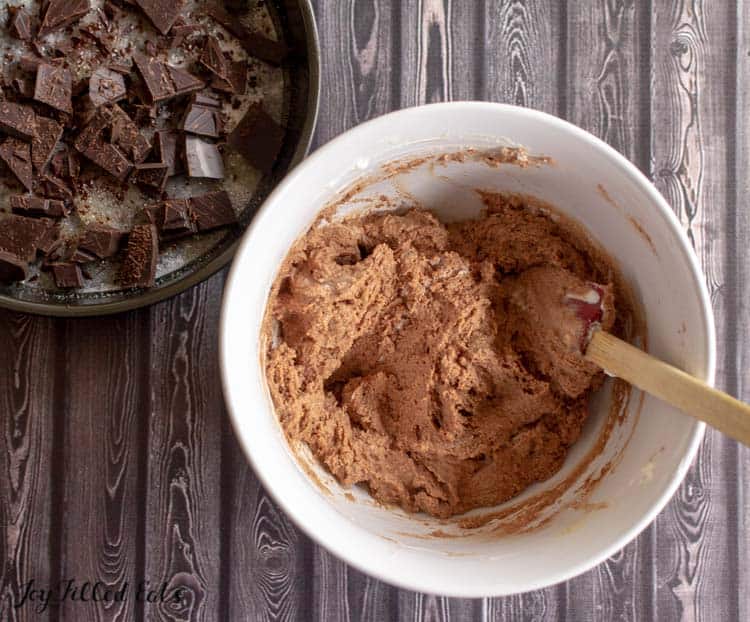 keto marble chocolate cake batter in mixing bowl with wooden spoon next to a bowl of broken chocolate pieces