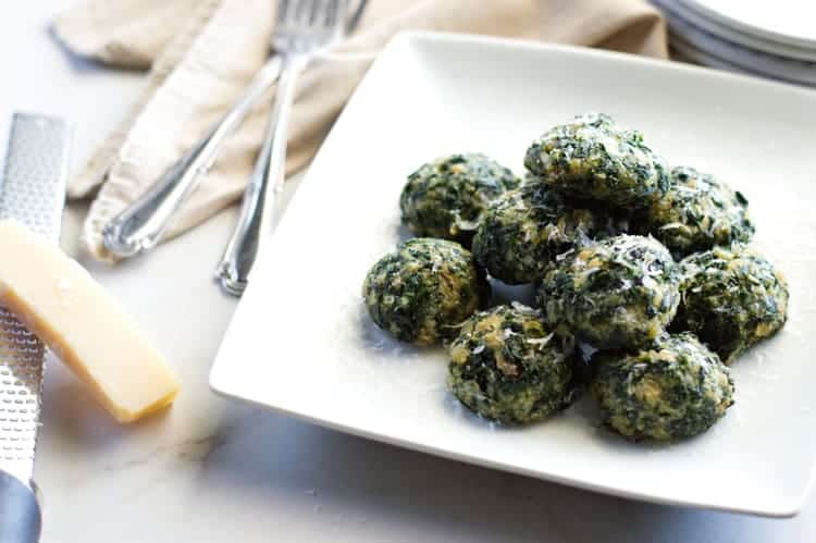 pile of spinach balls topped with Parmesan set on a table with forks, Parmesan rind and grater