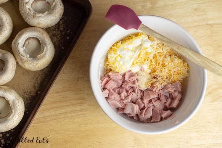 Ham and Cheese in white mixing bowl with spatula next to unstuffed mushrooms on sheet pan