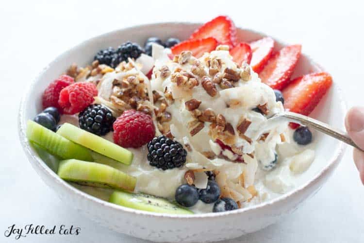 hand holding spoon lifting a bit of yogurt from bowl topped with various fruits and healthy granola