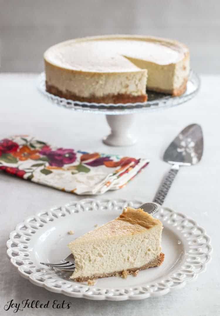 slice of ricotta cheesecake on a plate set in front of cake plate with remaining cheesecake
