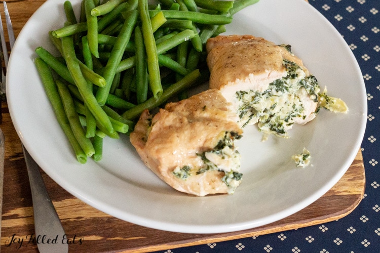 two halves of a keto stuffed chicken breast on white plate served with a side of green beans set on a cutting board next to a fork and knife