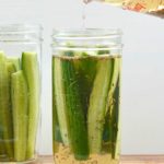 measuring cup pouring pickling liquid into mason jar of sliced mini cucumbers