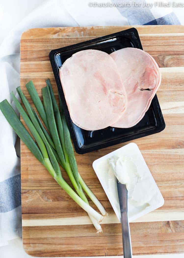 cutting board with sliced of ham, green onions and a knife with a block of cream cheese