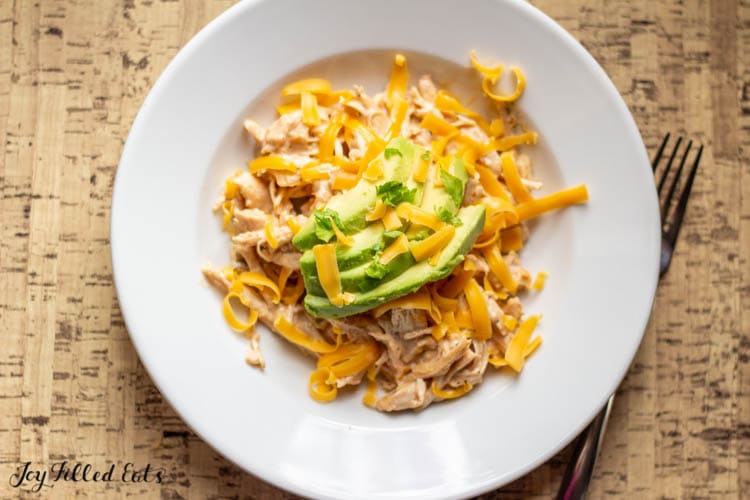 overhead view of creamy salsa chicken in a shallow white bowl topped with shredded cheese and avocado slices set next to a fork