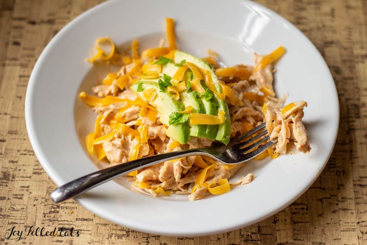 bite of creamy keto salsa chicken on a fork laying on the rim of a shallow white bowl filled with more creamy salsa chicken and topped with shredded cheese and sliced avocado