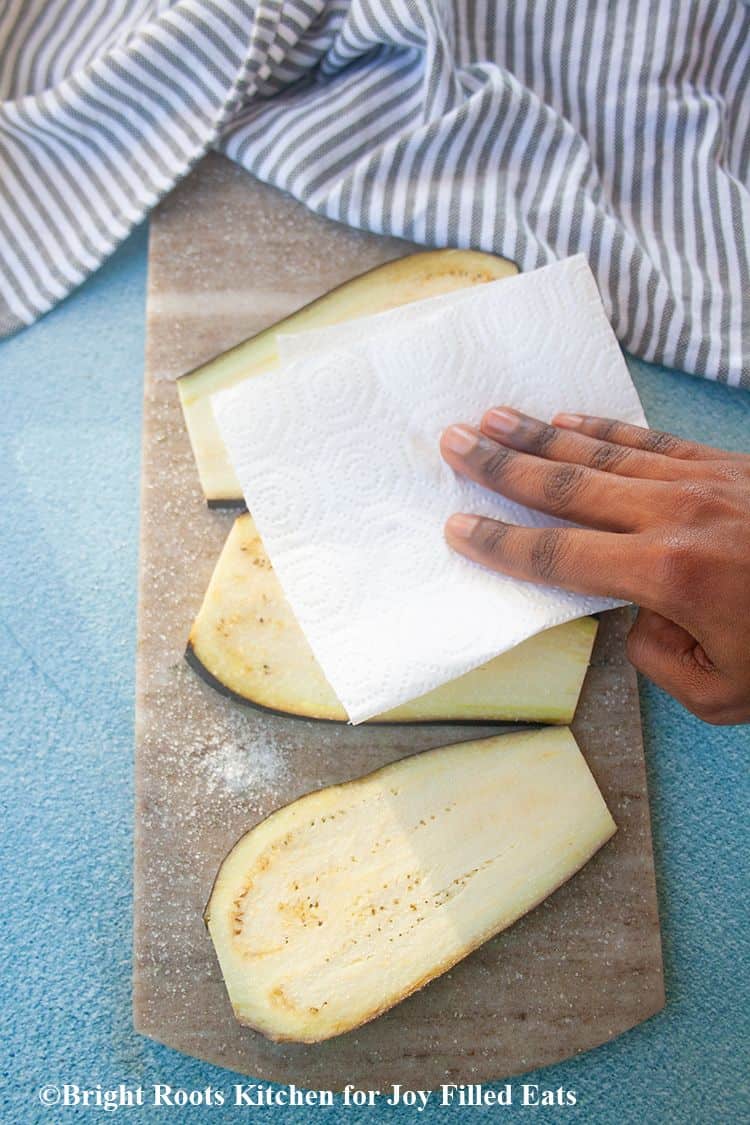 hand using paper towel to dry off eggplant slices on cutting board