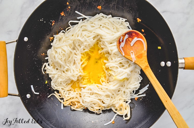 skillet and wooden spoon of pad thai noodles with egg poured into center