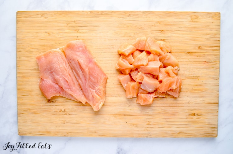 cutting board with raw chicken breast butterflied open and chicken cut into cubes