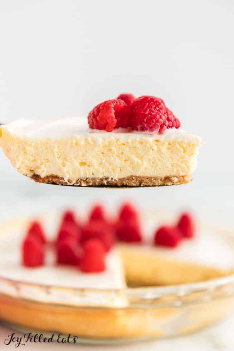 slice of keto lemon cheesecake topped with raspberries and a sour cream topping in front of remaining cheesecake in pie plate