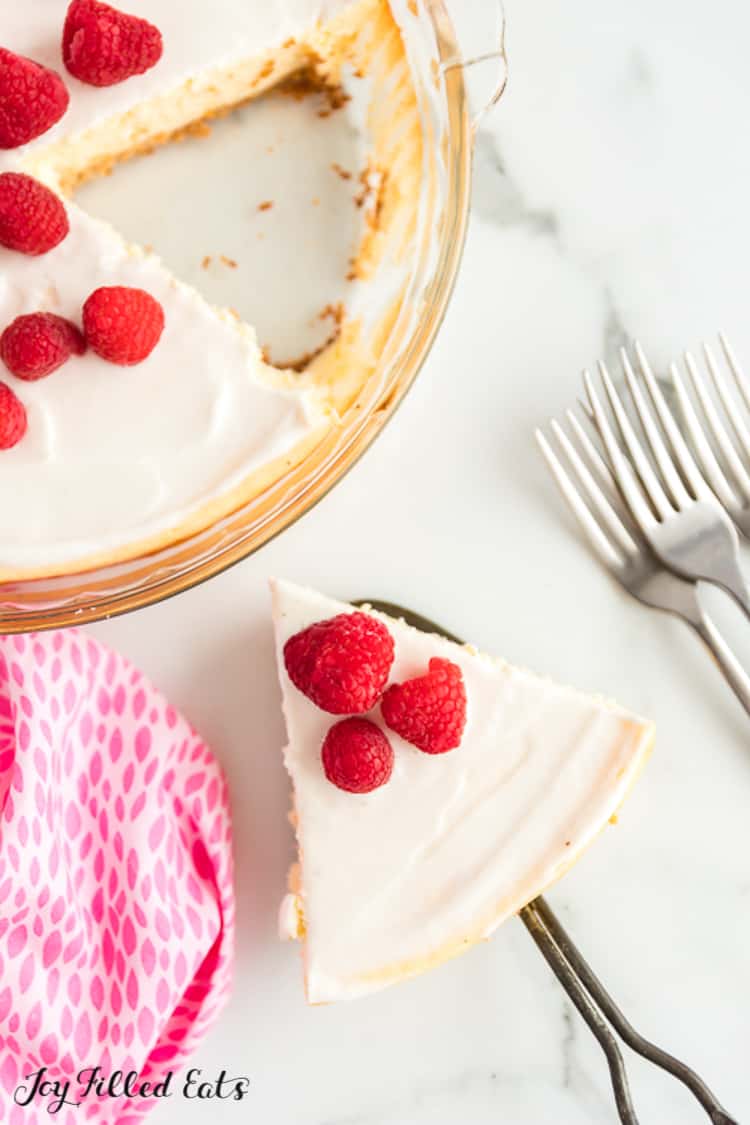 slice of keto lemon cheesecake topped with raspberries and a sour cream topping next to pie plate of remaining cheesecake and a handful of forks
