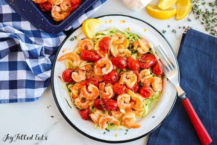 baked shrimp and garlic with cherry tomatoes in a bowl with fork next to sheet pan of more baked shrimp, garlic bulb and lemon wedges