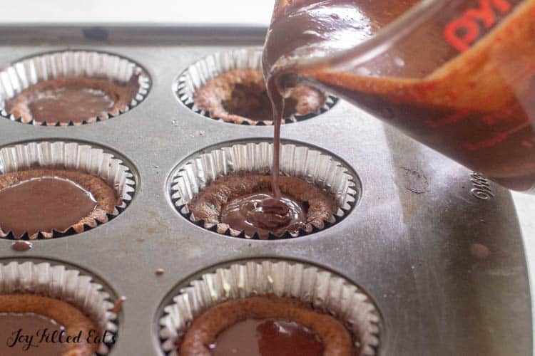 measuring cup of chocolate ganache filling being poured into chocolate chip cookie cups in a muffin tin