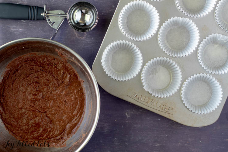mixing bowl of chocolate cupcake batter next to a sheet pan of empty cupcake wrappers and an ice cream scoop