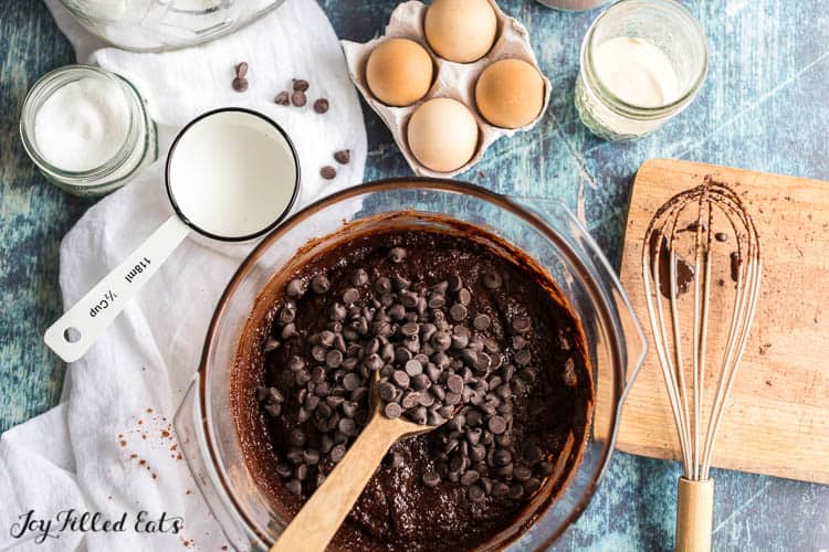 chocolate chips being mixed into large bowl of brownie batter with wooden spoon surrounded by ingredients and kitchen utensils
