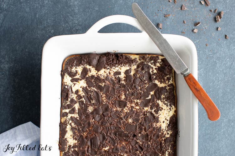 keto chocolate cake bars with cheesecake swirl and chocolate shavings topping in a baking dish with knife resting on rim