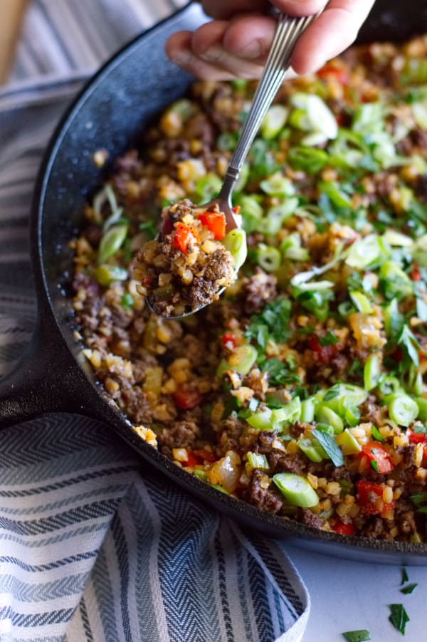 hand holding spoon with scoop of dirty cauliflower rice on it above skillet full of cauliflower rice