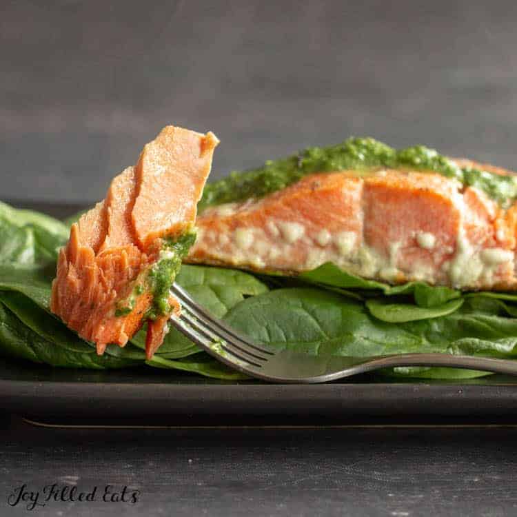 fork with salmon piece resting on plate of baby spinach with a salmon filet topped with pesto close up